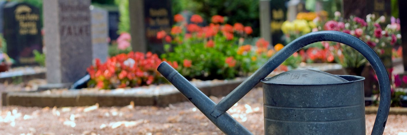 Gießkanne aus Metall auf einem Friedhofsweg vor Gräbern mit Grabsteinen