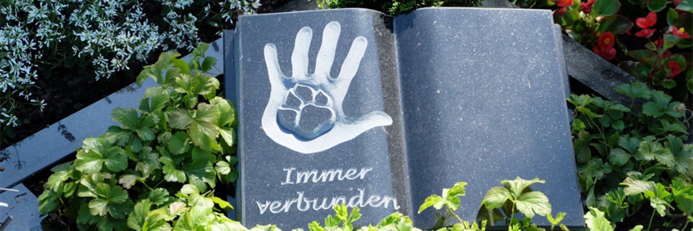 Dunkler Grabstein in Form eines aufgeklappten Buches mit Symbol aus Tierpfote und Hand eines Menschen
